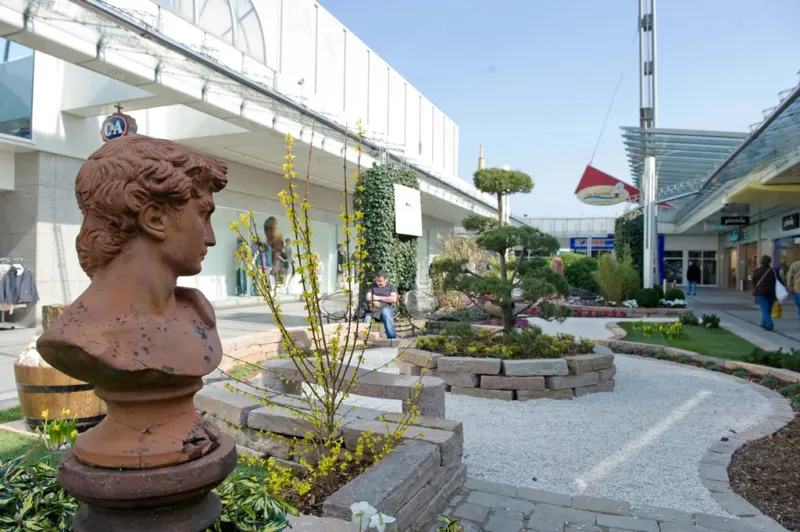 Antike Terrakotta-Büste auf einem Sockel, umgeben von Gräsern und Stauden im Schaugarten
